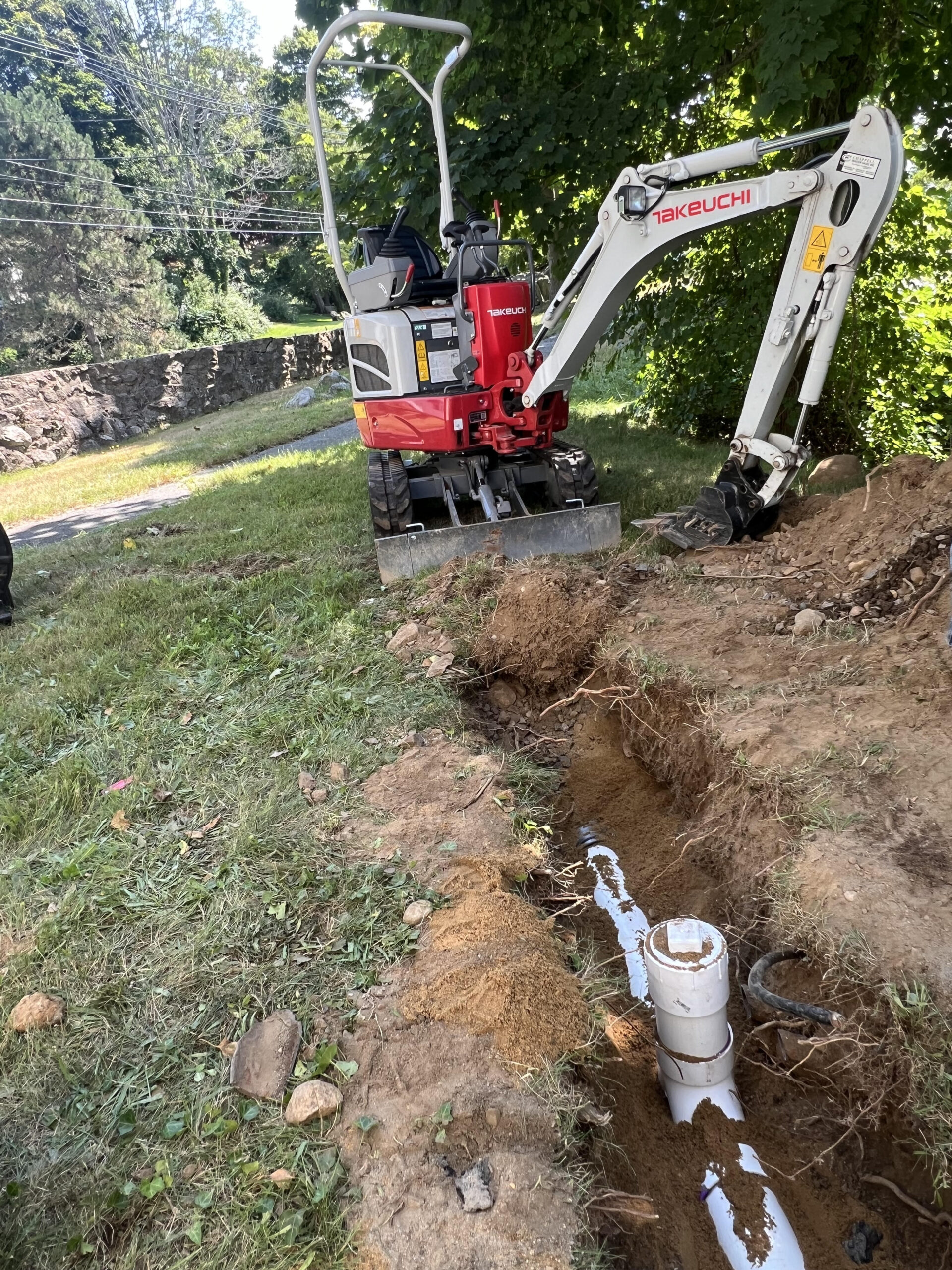 Photo of an excavator digging a trench for sewer repairs, with newly installed sewer line visible in the trench.