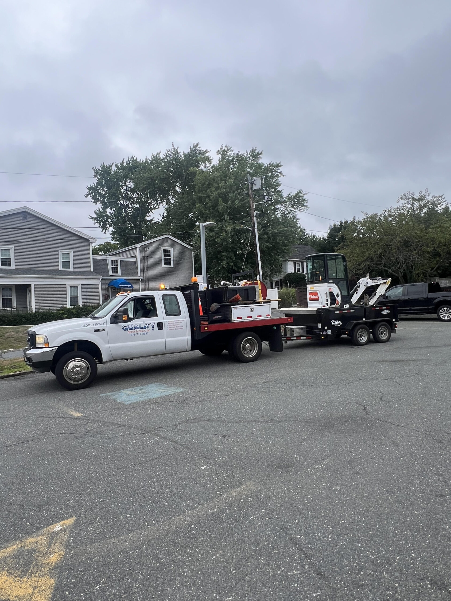 Photo of a Quality Sewer & Drain, Inc. service truck towing an excavator on a trailer, parked in a residential area.